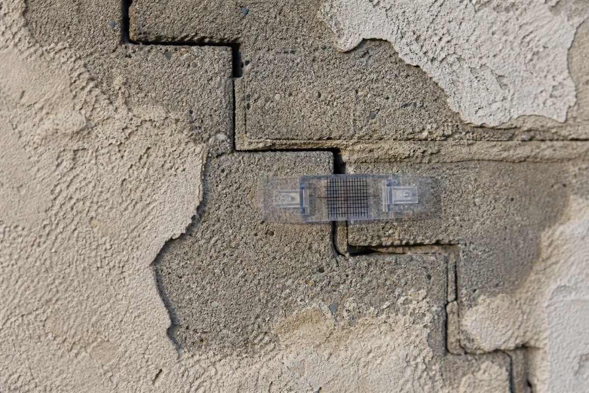 Fissure en escalier suivant les joints d&rsquo;un mur en parpaings enduit, avec un fissuromètre gradué collé en travers